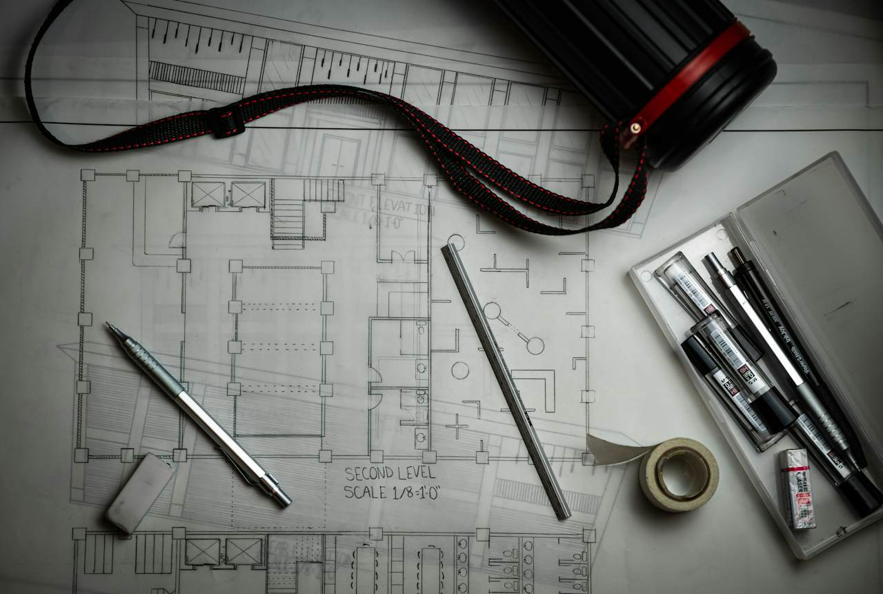 A pencil-drawn architectural floor plan with a mechanical pencil, an eraser, a ruler and a case of pens, representing a plan that is about to be revised